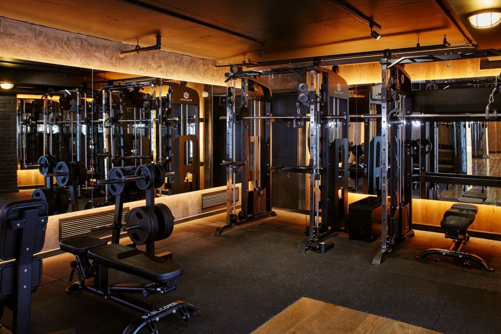 A high-end strength training area equipped with multiple functional cable machines and a weight bench. The room is lit with warm, moody orange lighting against dark ceilings and mirrored walls. The floor is covered in professional black gym matting.