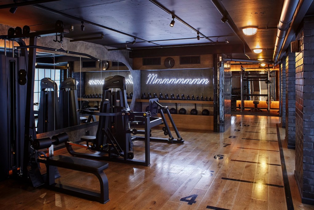 A sleek weight training room featuring various resistance machines and a row of kettlebells and medicine balls on a wooden shelf. A white neon sign that reads "Mmmmmmmm" glows on the concrete wall. The wood floor includes numbered markers for guided workouts.