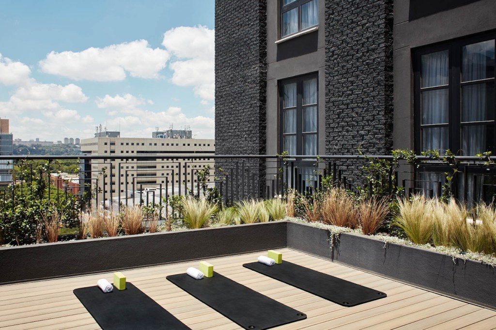 An outdoor yoga terrace on a rooftop featuring three black yoga mats neatly laid out with blocks and rolled white towels. The deck is made of light wood planks, surrounded by a dark grey planter wall with ornamental grasses and a metal railing. The background shows an urban landscape under a bright, partly cloudy sky.