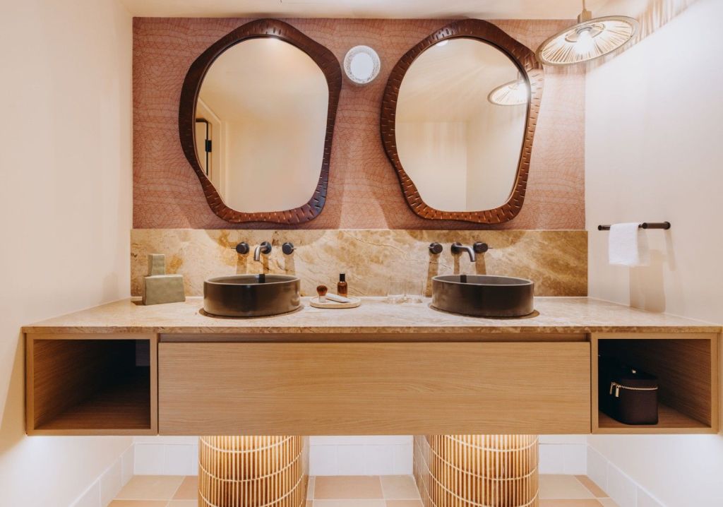 Warm-toned bathroom with a floating vanity, stone countertop, two faucets and two large round mirror.