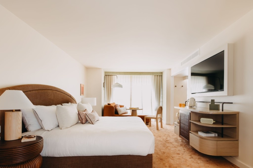 Warm bedroom with a king bed, bedside lamps on wooden stools, and a cozy sitting area by a green accent wall featuring a leather chaise, round table, and chair.