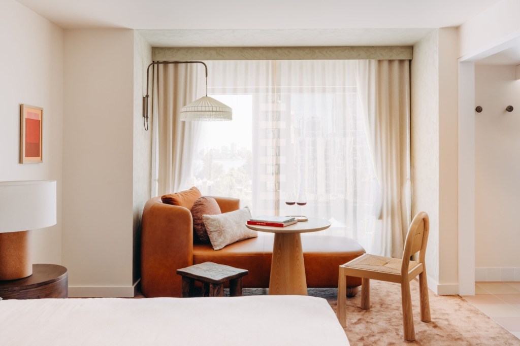 Warm bedroom with a king bed, bedside lamps on wooden stools, and a cozy sitting area by a green accent wall featuring a leather chaise, round table, and chair.