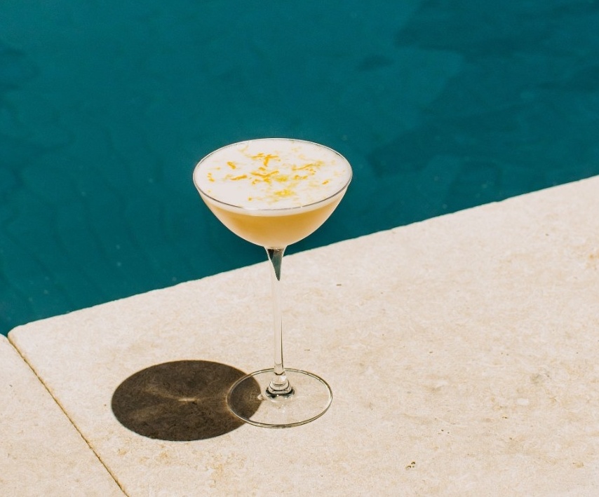 Poolside scene with a coupe glass cocktail topped with foam and citrus zest ,set against pool water.