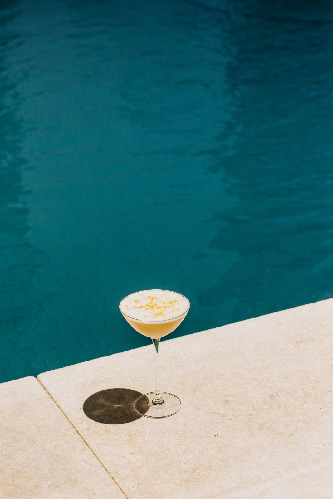 Poolside scene with a coupe glass cocktail topped with foam and citrus zest ,set against pool water.