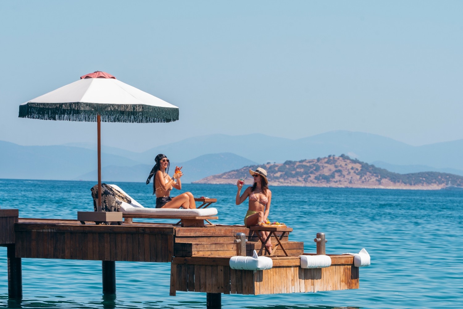 Two women sitting by the beach on a wooden deck enjoying their cocktails