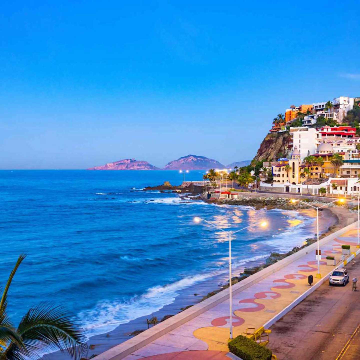 Cars drive along a road beside the ocean and a Mazatlan beach at dusk