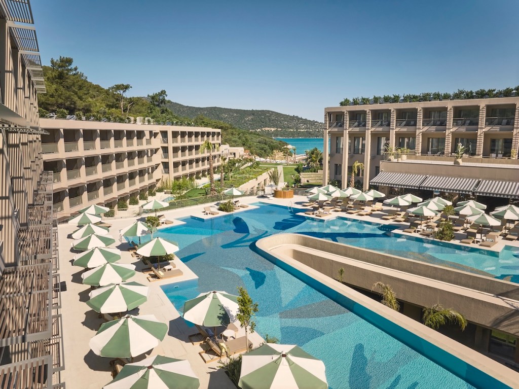 Overhead shot of a pool with interesting design at the bottom and white and green umbrellas over lounge chairs with a forest and sea in the far distance