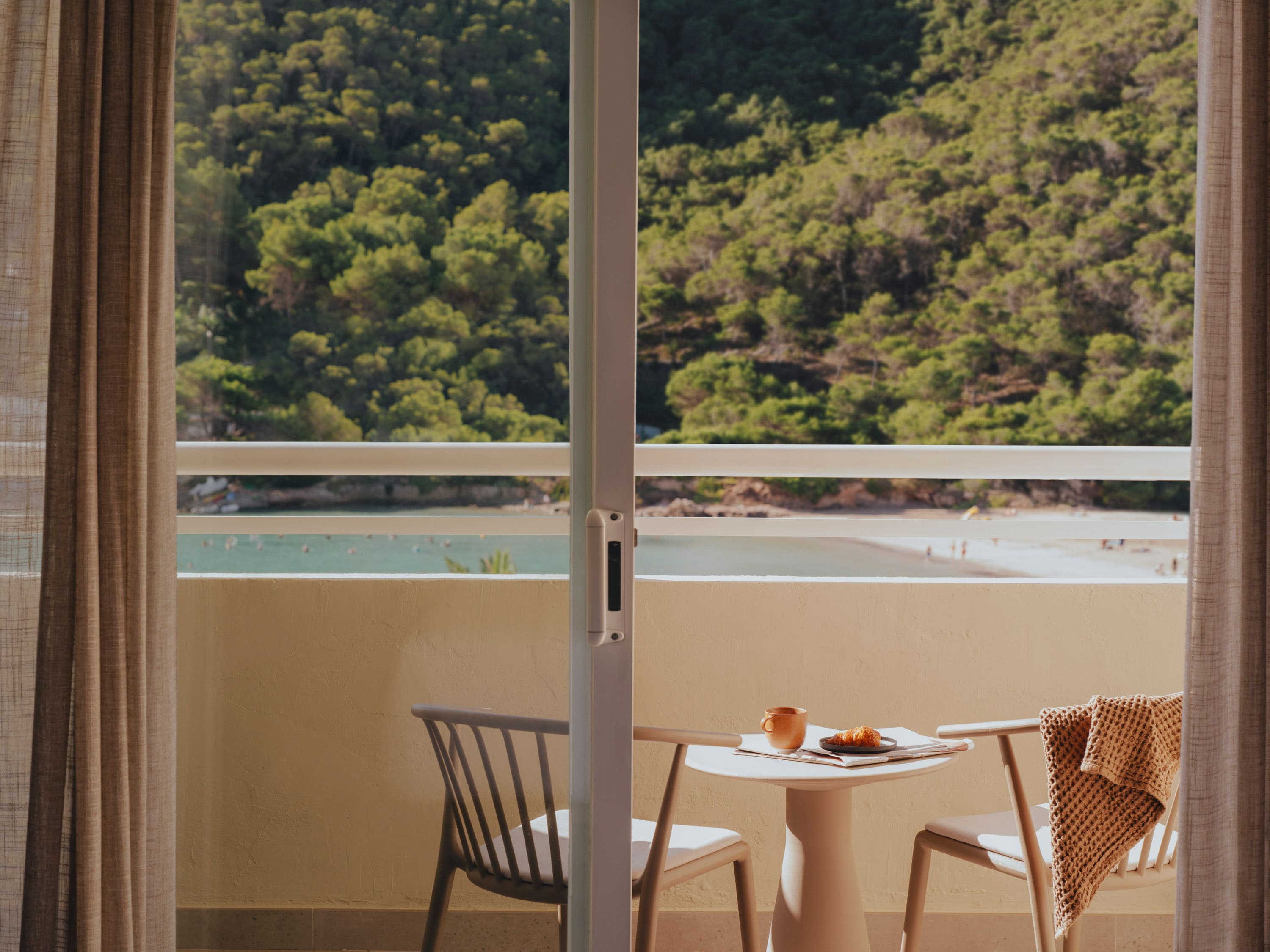 two chairs and a small table in a balcony with the view of the sea and a cliff with greenery on it