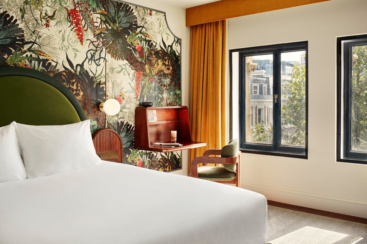 Sunlit bedroom with white double bed, a desk with green chair to the right and bright wallpaper with views of London from the window