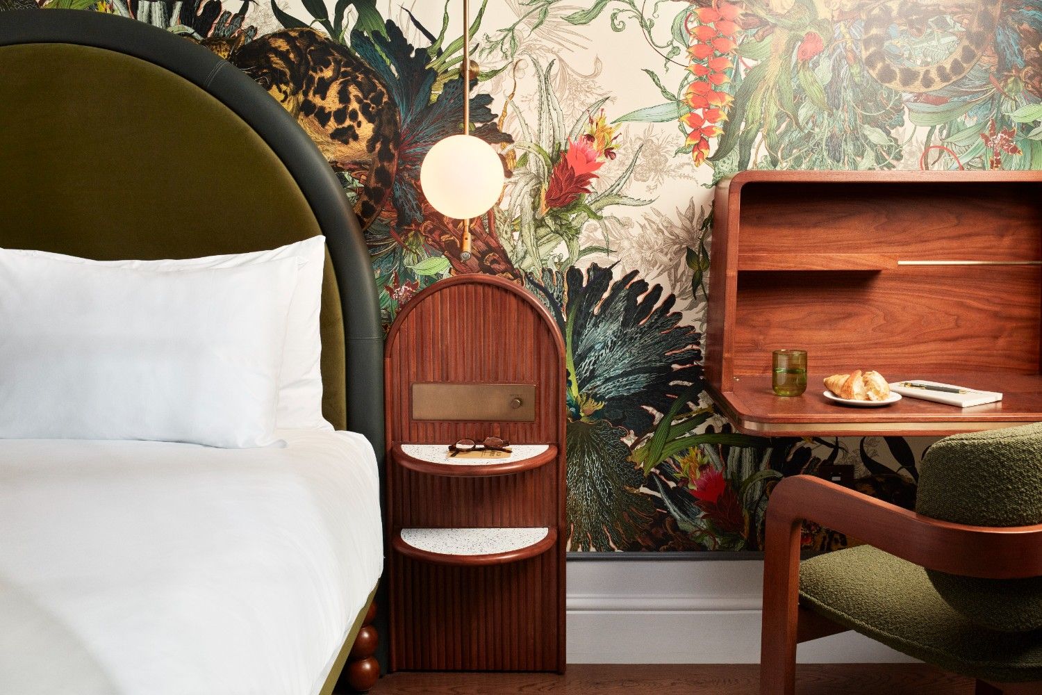 One side of a double bed showing the white bedding and pillow, with a dark wooden side table and desk next to it