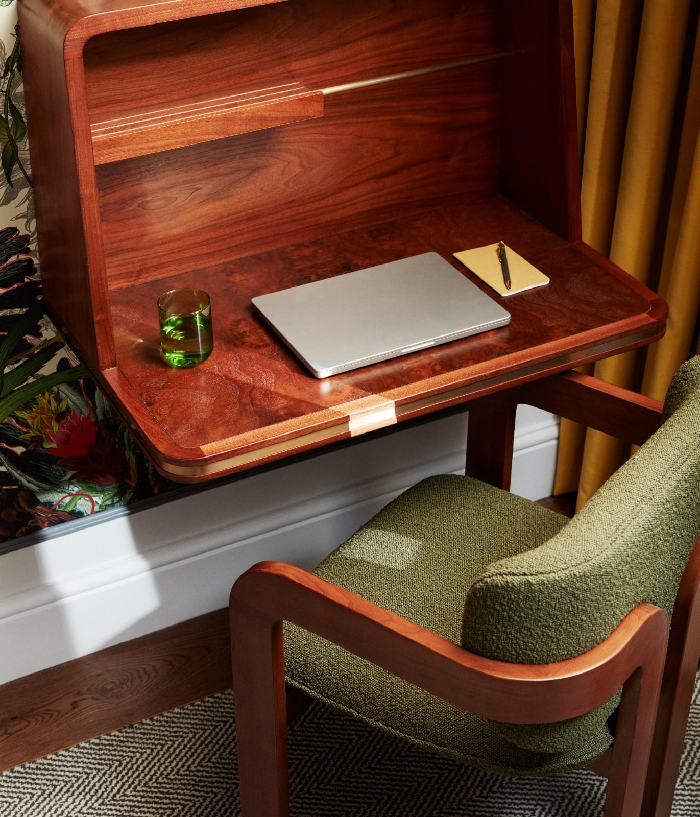 A dark green desk chair with a wooden arm, pulled out of mahogany desk with writing materials on it