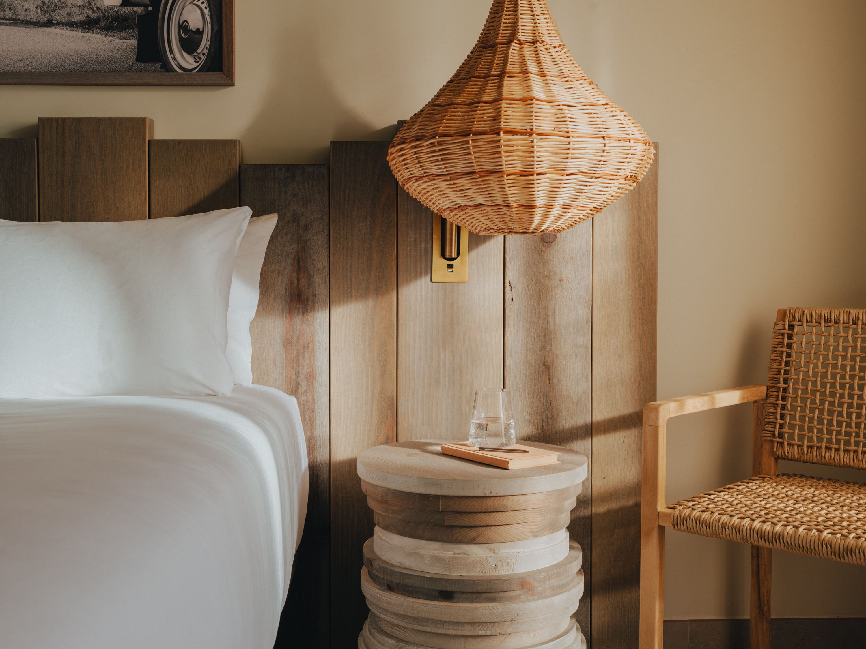 close up of bedside table with glass of water on it and bed next to it and ceiling light hanging closely