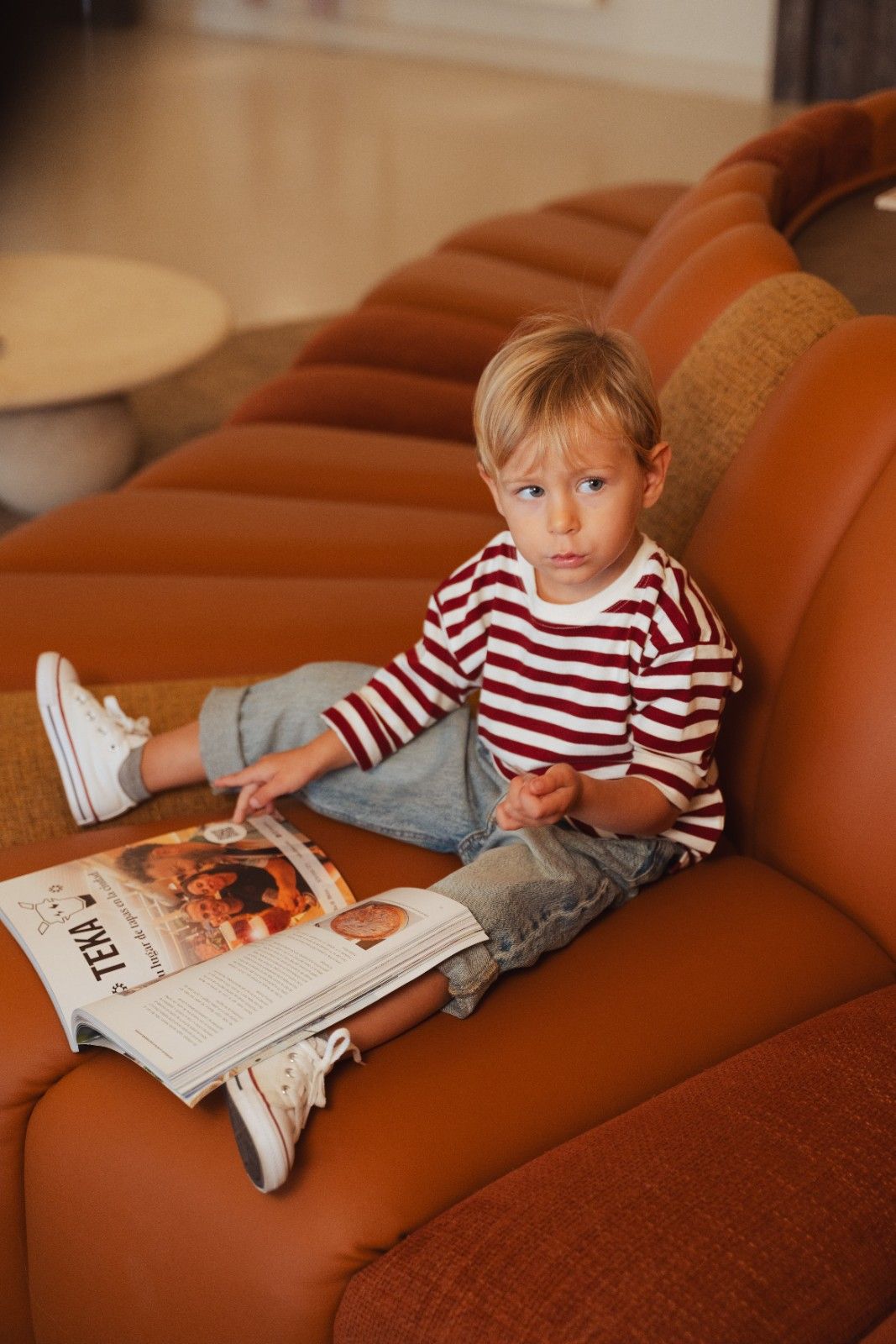 Blonde child dressed in white and red striped top and jeans on red sofa