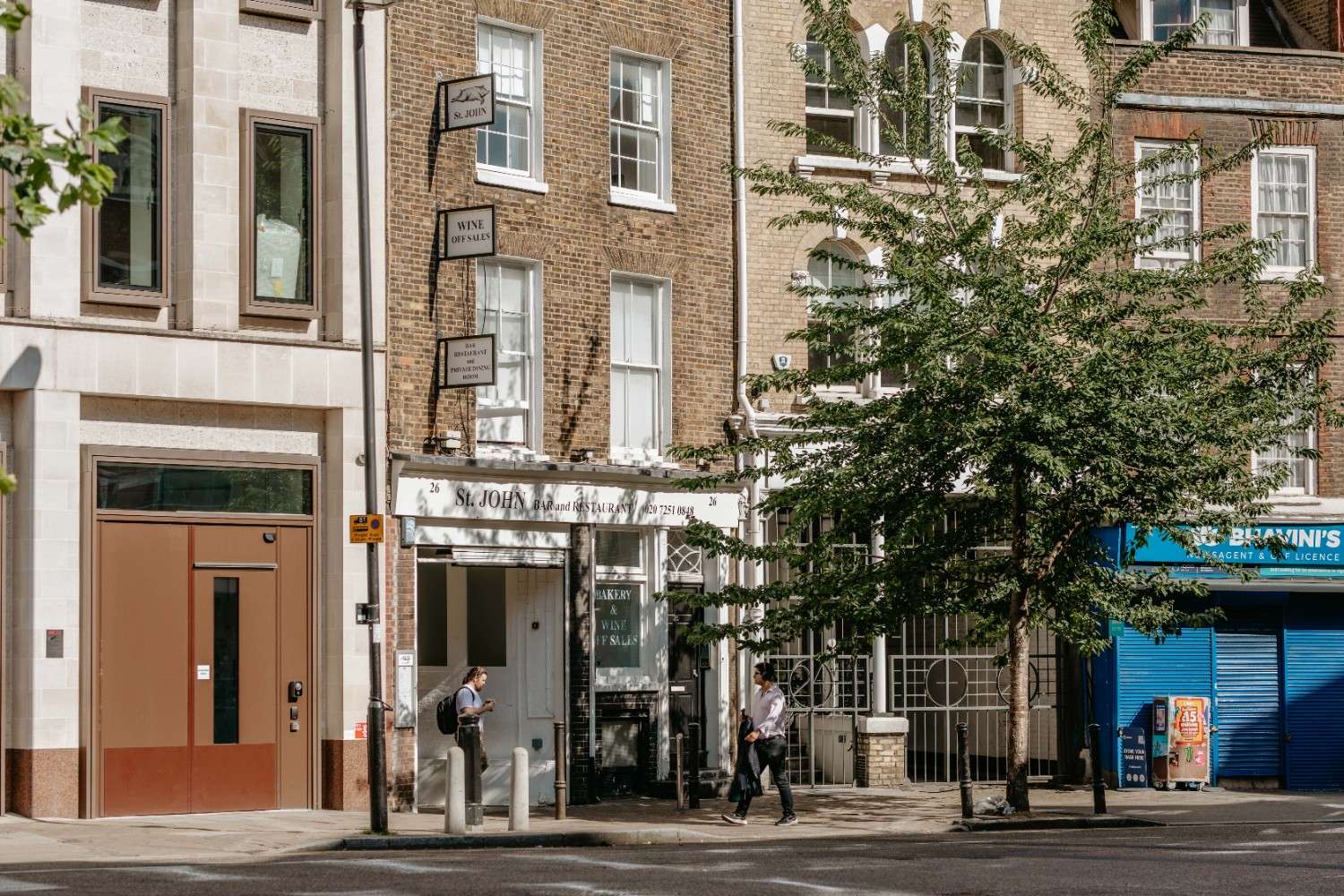 Farringdon neighbourhood in the sun, showing a row of shops and cafes with a large tree outside and people strolling