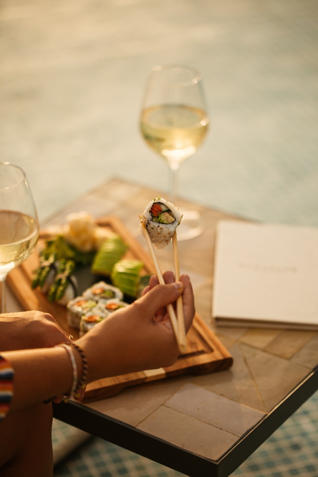 a plate of sushi and a glass of wine in the background with someone using chopstick to eat the sushi