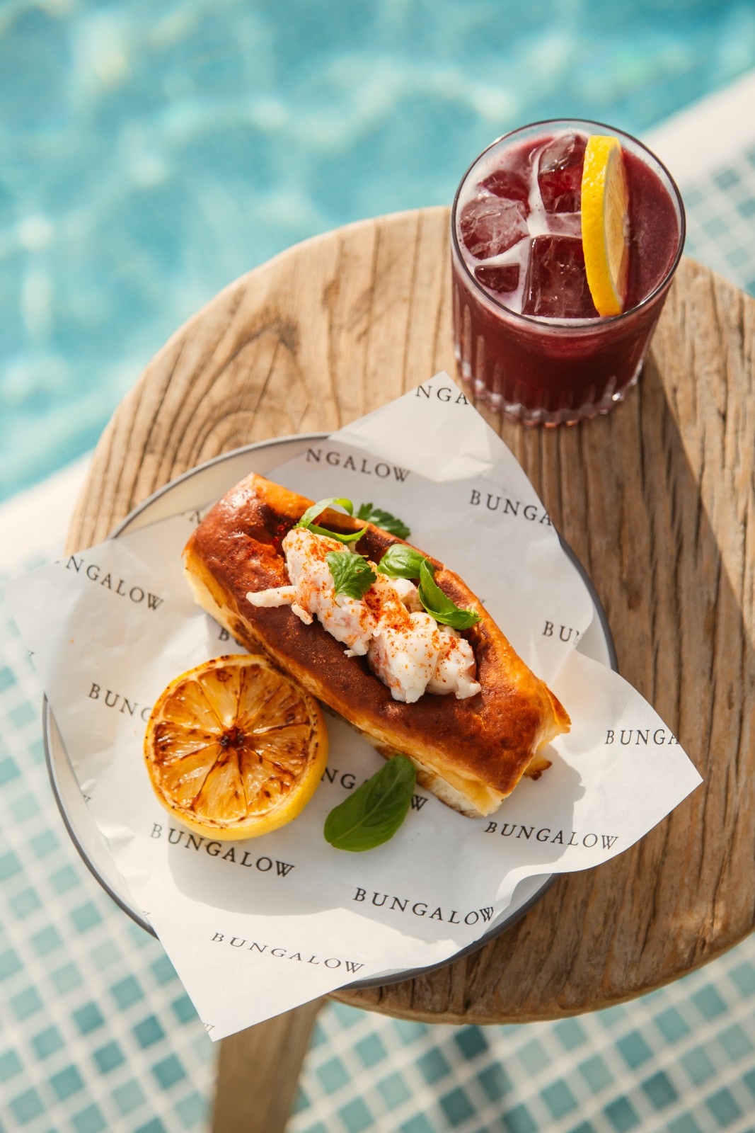 a lobster roll and a red cocktail on a table by the pool