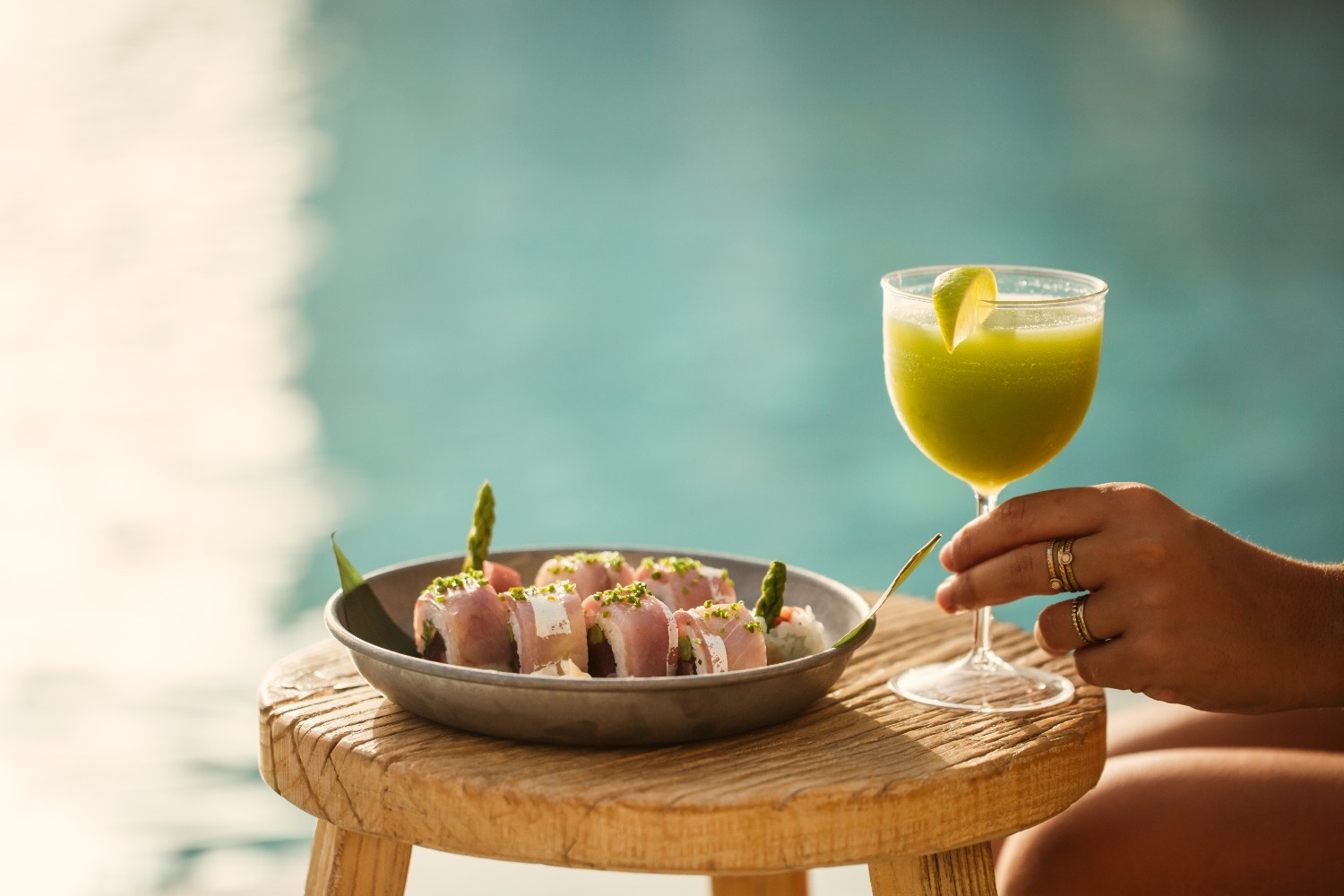 a plate with sushi on a table with someone holding a green drink next to it with the pool in the background