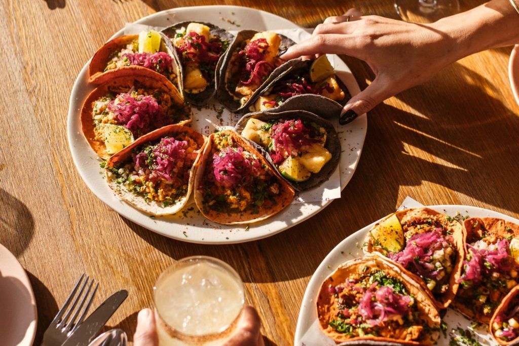 Multiple colourful tacos on a plate with someones hand showing as they are grabbing a taco