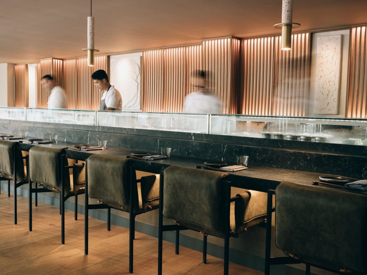 bar of a restaurant with empty high chairs against it and some chefs behind it working