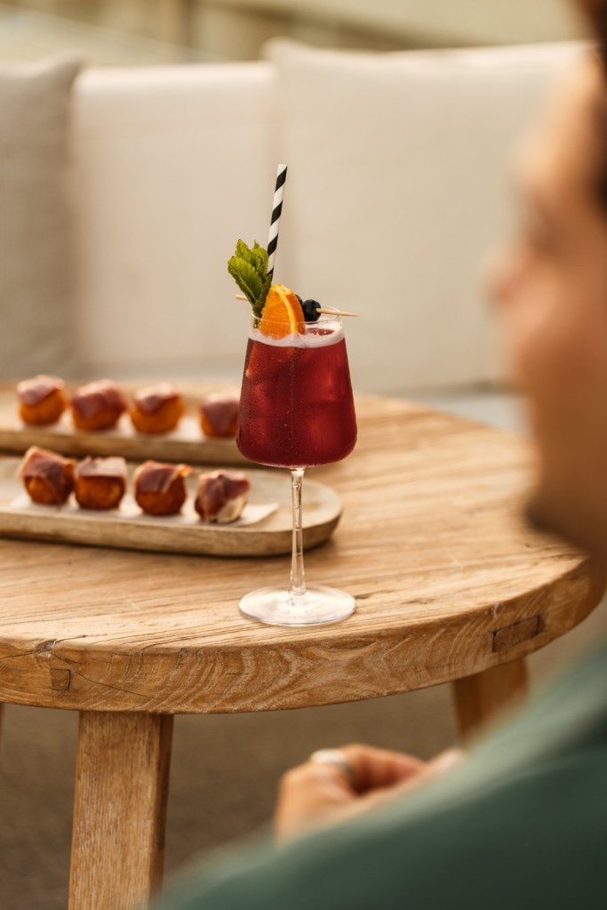 Une belle boisson rouge avec une tranche d'orange et quelques morceaux verts sur une table, avec une assiette de bouchées derrière.