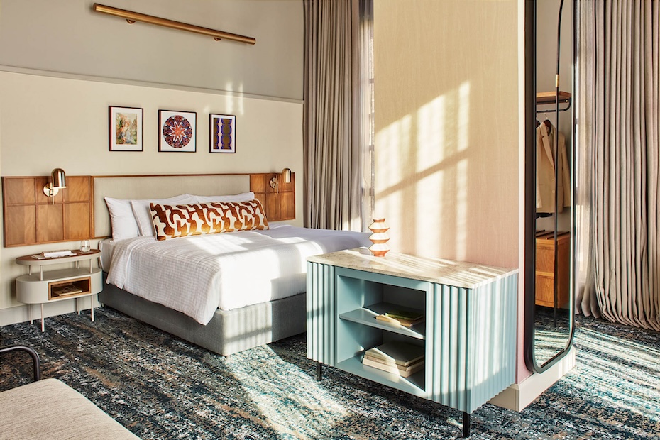 A full view of a stylish hotel bedroom showing a plush bed with a wood-paneled headboard and a long decorative bolster pillow. A light-blue fluted cabinet serves as a room divider, with a full-length mirror reflecting the open closet area.