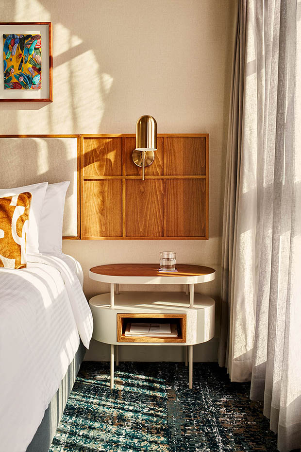 A stylish two-tier oval bedside table in white and wood holding a glass of water. Above it is a brass pill-shaped sconce mounted on a wood wall panel, set against light-colored wallpaper and sheer curtains.
