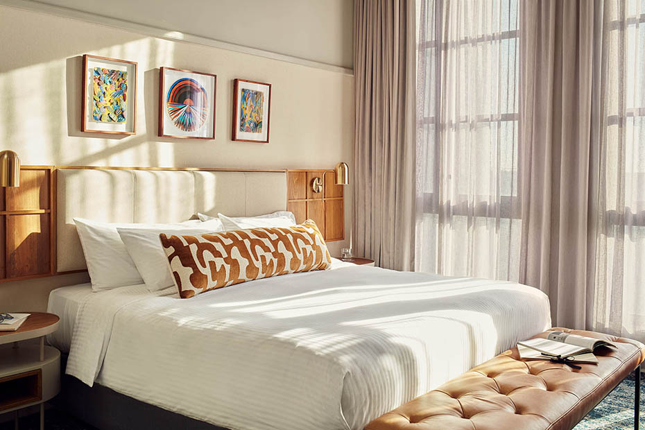 A wide shot of a bright hotel room featuring a king-sized bed with crisp white bedding, a tan leather tufted bench at the foot, and three framed art pieces above the headboard. Large windows with sheer curtains let in soft, natural light.