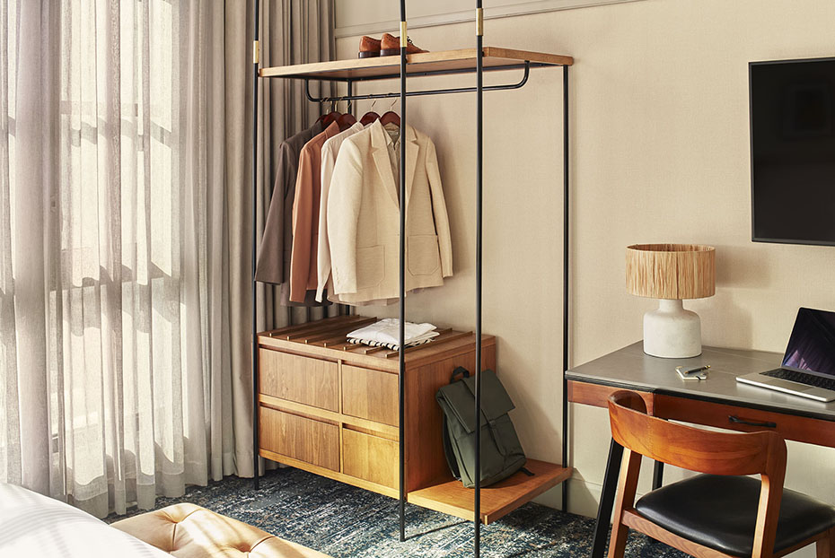 An open-concept industrial wardrobe with wood drawers and a black metal frame holding blazers and a backpack. To the right is a dark wood desk with a mid-century modern chair and a lamp with a fringed shade.