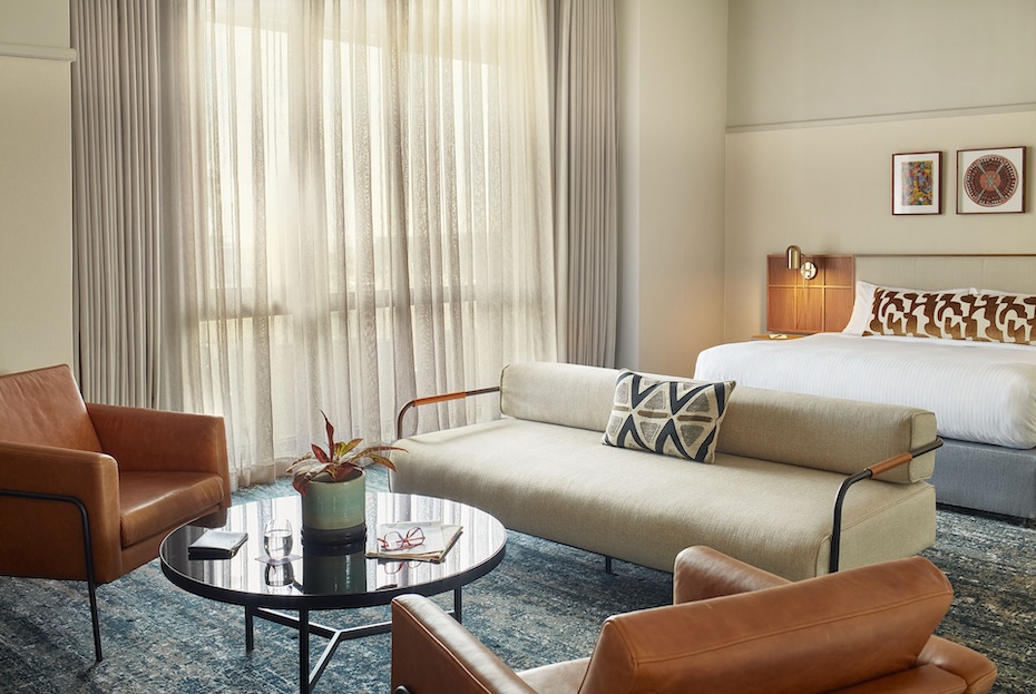 A spacious hotel suite living area with floor-to-ceiling sheer curtains filtering soft sunlight. The room includes two tan leather armchairs, a low-profile cream sofa with a geometric print pillow, and a black circular coffee table set on a textured blue and grey area rug.