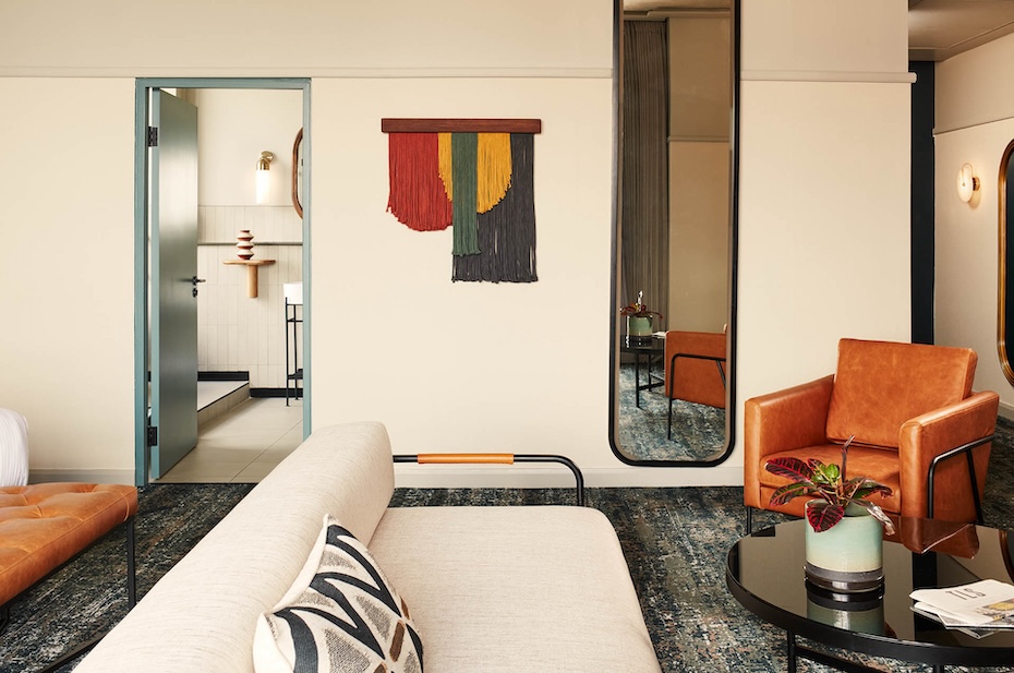 An interior view of the suite showing a transition from the living area to the bathroom. A colorful textile wall hanging in red, green, and yellow adds a pop of color next to a tall, thin arched floor mirror. The open door reveals the modern tiled bathroom in the background.