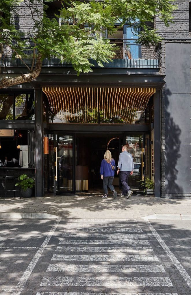A modern architectural entrance of a hotel with dark grey brick walls and large glass doors. Above the entrance, a decorative wooden slat ceiling with warm lighting creates a wave-like effect. A couple is walking toward the entrance from a paved street with a white-painted pedestrian crossing in the foreground.