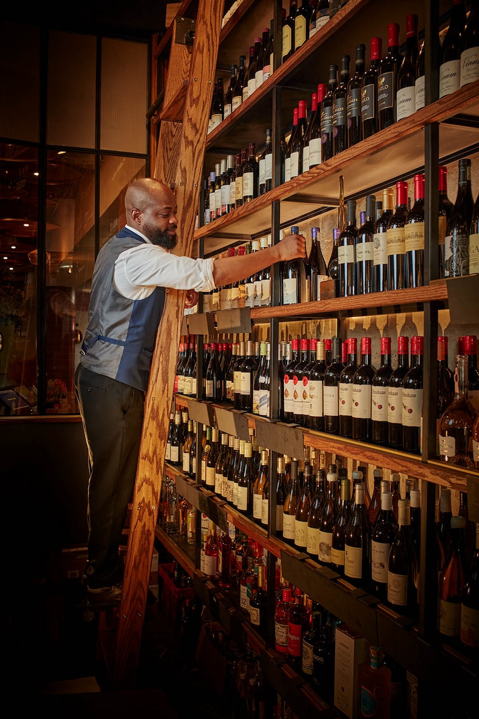 A professional sommelier or wine steward standing on a wooden library ladder to reach the upper shelves of an expansive, floor-to-ceiling wine cellar. He is carefully selecting a bottle from rows of red and white wines. The lighting is warm and dramatic, highlighting the extensive collection.