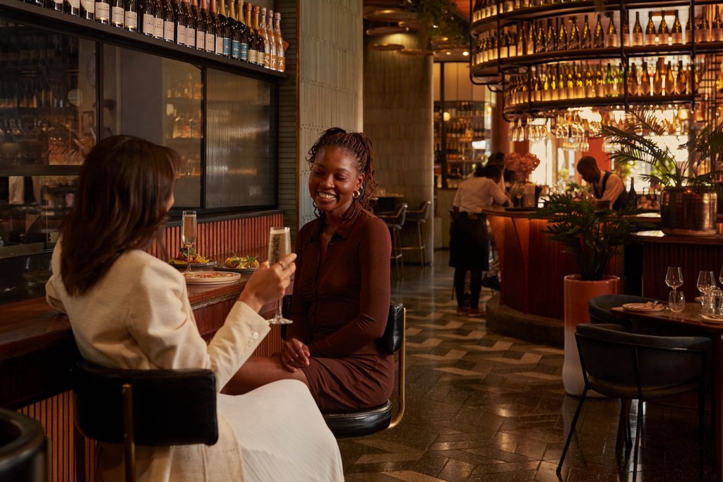 Two women enjoying drinks at a long bar counter inside a luxury restaurant. One woman, seen from the back in a white blazer, raises a champagne flute in a toast toward her friend, who is laughing. The background shows an expansive, well-lit dining area with a circular bar and professional staff.