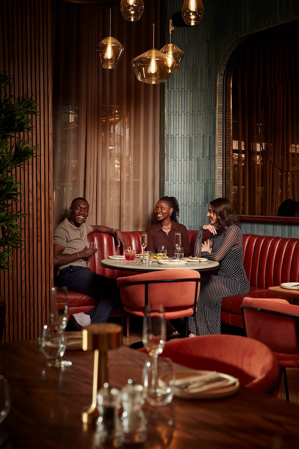 A group of three friends sharing a meal and drinks in a cozy, upscale restaurant booth. They are seated on a curved red leather banquette at a round marble table. Warm, amber-toned glass pendant lights hang above them, casting a glow against a backdrop of teal vertical tiles and wooden slats.
