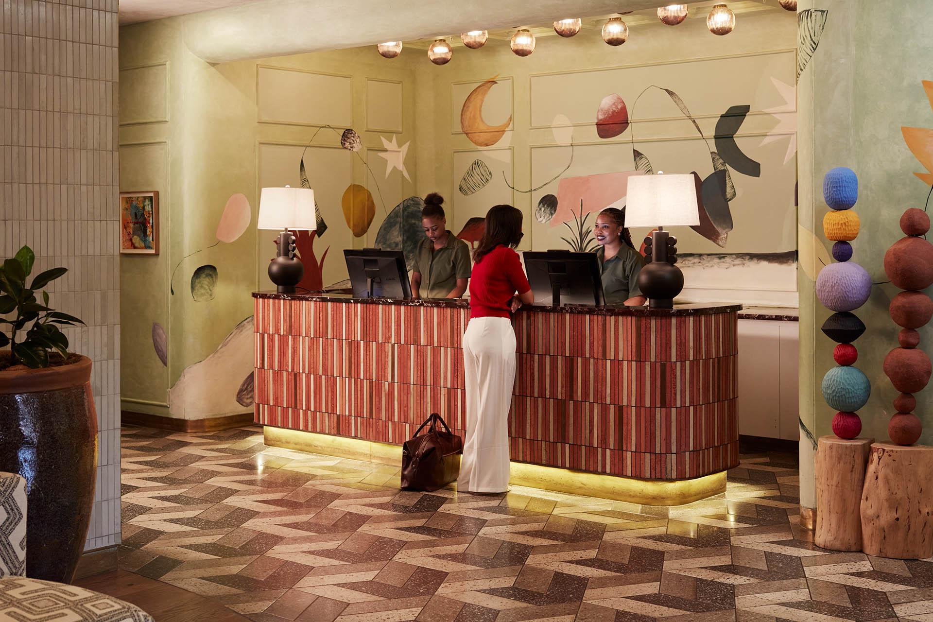The hotel reception area featuring a wood-slatted desk with gold under-lighting. The back wall is decorated with a colorful, abstract mural, and the space is accented by unique totem-like sculptures and modern lamps.