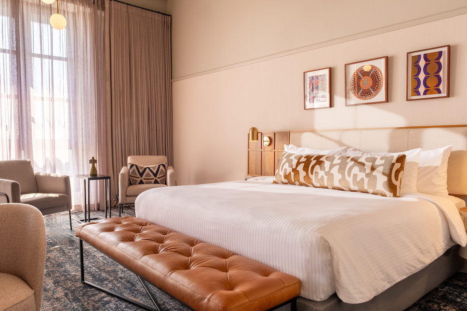 A wide view of a modern hotel suite featuring a king-size bed with white striped linens and a long patterned bolster pillow. A tan tufted leather bench sits at the foot of the bed, and two grey armchairs flank a small side table by a large window with sheer curtains.