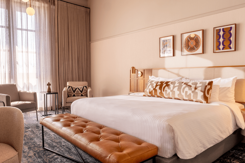 A wide view of a modern hotel suite featuring a king-size bed with white striped linens and a long patterned bolster pillow. A tan tufted leather bench sits at the foot of the bed, and two grey armchairs flank a small side table by a large window with sheer curtains.