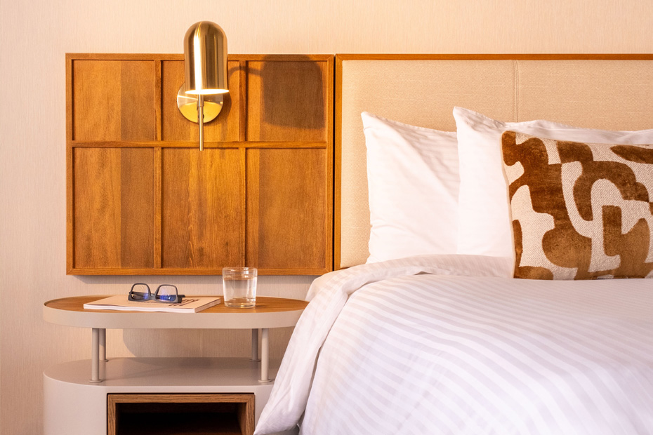 A close-up of the bedside lighting and headboard area, showing a sleek brass reading lamp mounted to a wood-paneled wall section. The bed is made with white striped linens and features a plush tan-and-cream patterned bolster pillow.