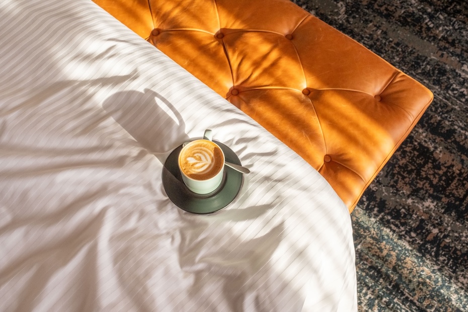A top-down lifestyle shot of a cup of latte with foam art resting on a white striped duvet. The warm sunlight casts shadows across the bed and a portion of the tan tufted leather bench.