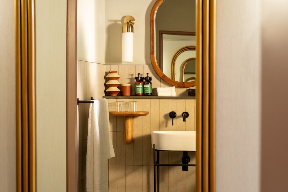 A glimpse into a modern bathroom through a doorway, showing a white vessel sink with black matte fixtures, a round wooden-framed mirror, and a small wooden shelf holding drinking glasses.