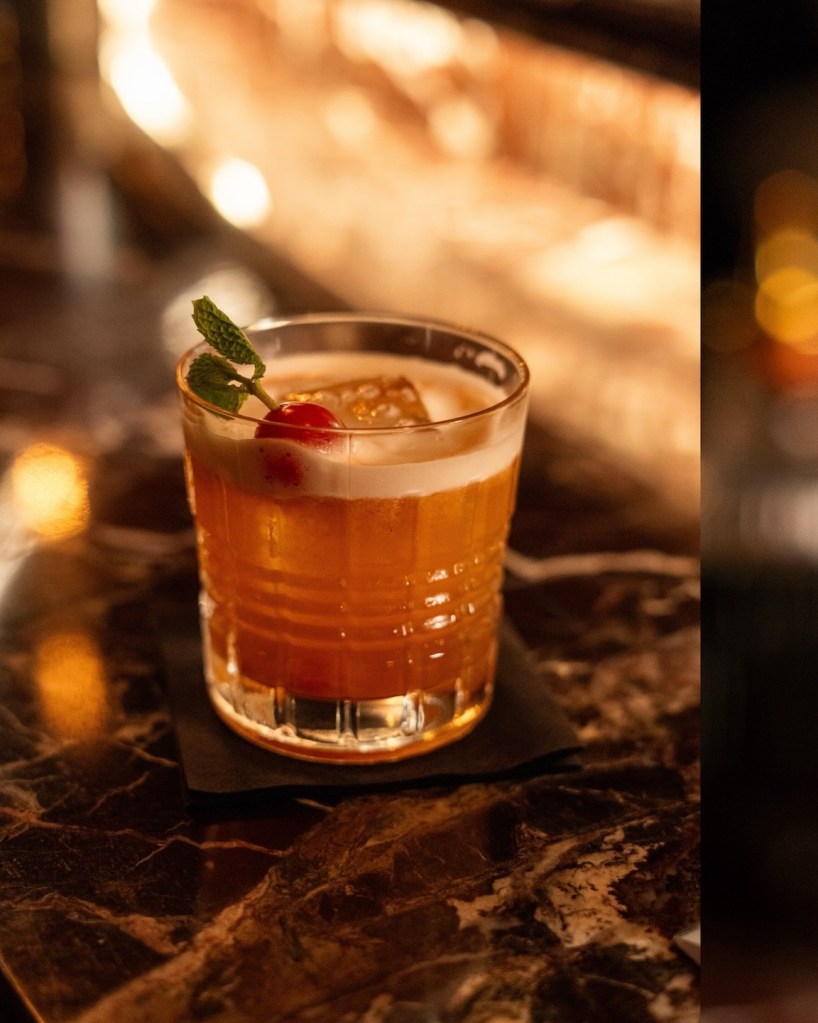A short, ribbed crystal glass containing an amber-colored cocktail over a large ice cube, garnished with a red cherry and a sprig of fresh mint. The drink sits on a black napkin atop a dark marble surface with warm, bokeh lighting in the background.