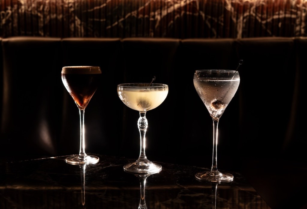 Three different cocktails in elegant glassware—a dark espresso martini, a pale yellow coupe, and a clear martini with an olive—lined up on a dark marble surface against a black background.