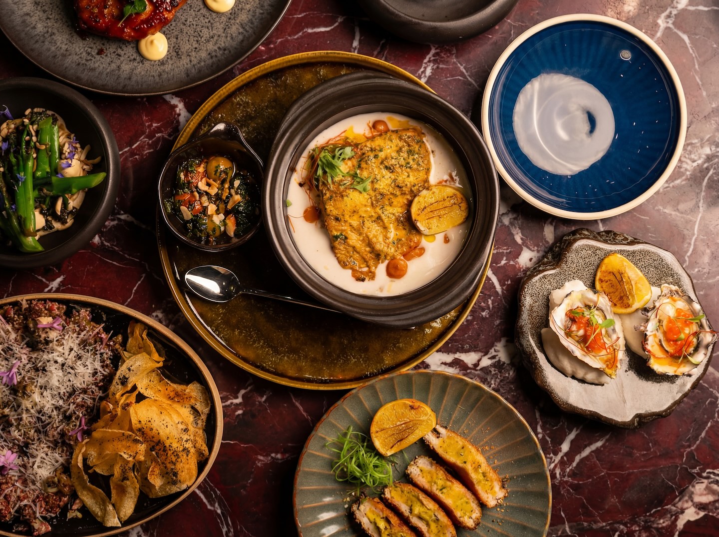 An overhead spread of various small plates on a reddish-marble table, including oysters with roe, a breaded cutlet dish, seared meat with chips, and a central bowl of fish in a creamy sauce.