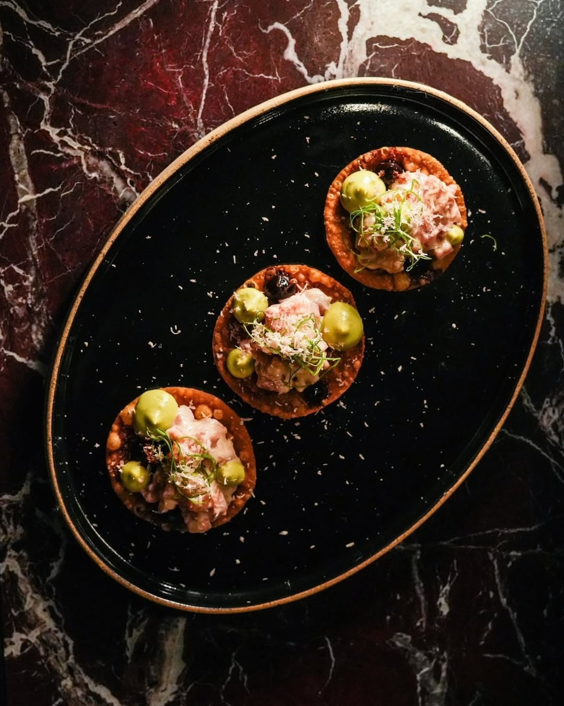 Three round tostada-style appetizers arranged on a black oval plate. Each is topped with a creamy pink mixture, dollops of green sauce, and fine garnishes, set against a dark marble background.