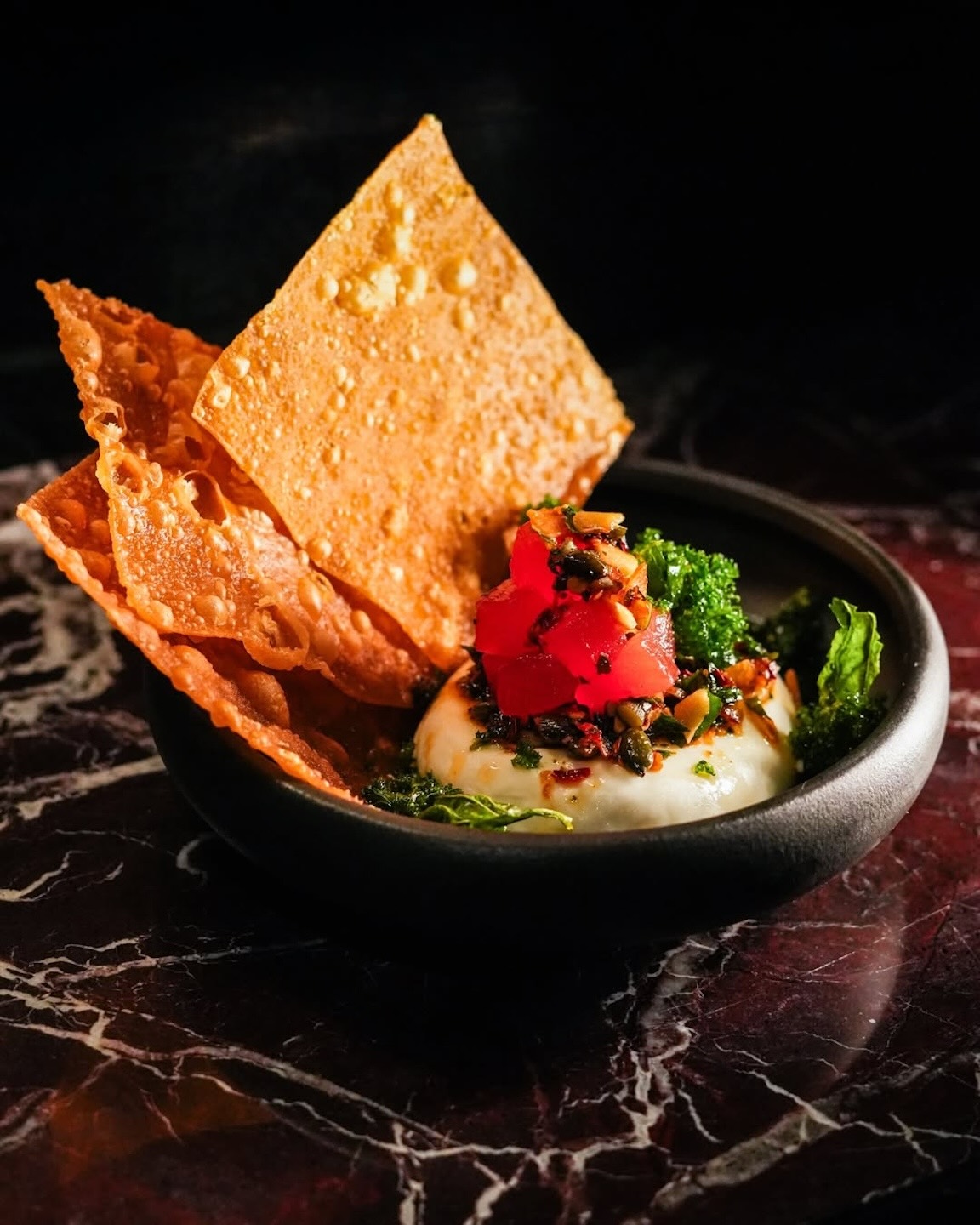 A small black bowl containing a white creamy base topped with red diced tuna and fresh greens, served with large, crispy golden crackers standing upright.