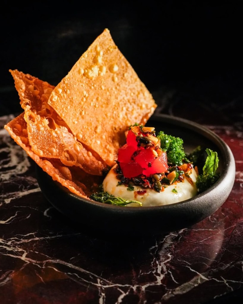 A small black bowl containing a white creamy base topped with red diced tuna and fresh greens, served with large, crispy golden crackers standing upright.