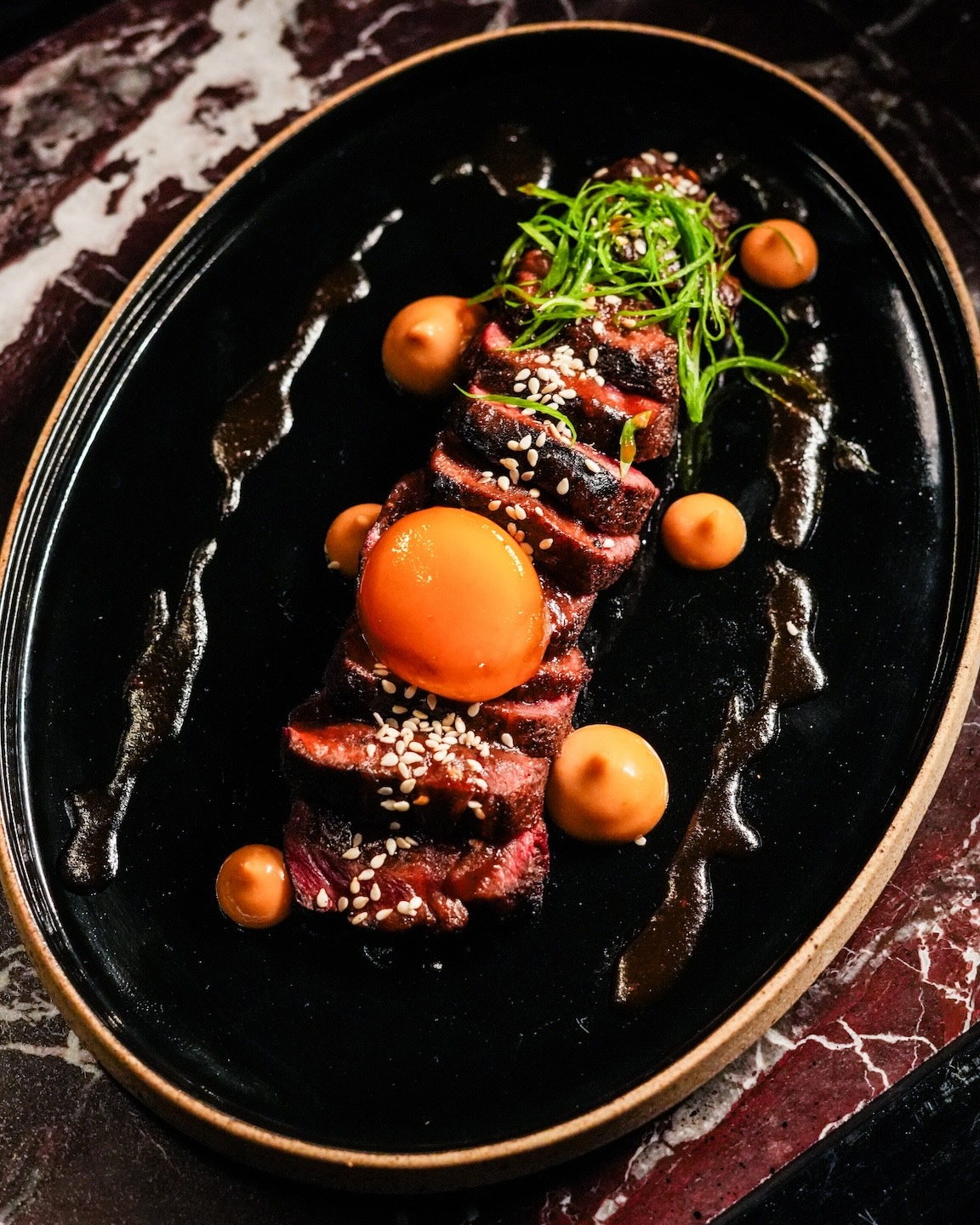 Sliced seared beef tataki arranged on a black oval plate, topped with sesame seeds, micro-greens, and a bright orange egg yolk. The dish is finished with dots of creamy sauce and a dark glaze, set on a dark marble tabletop.