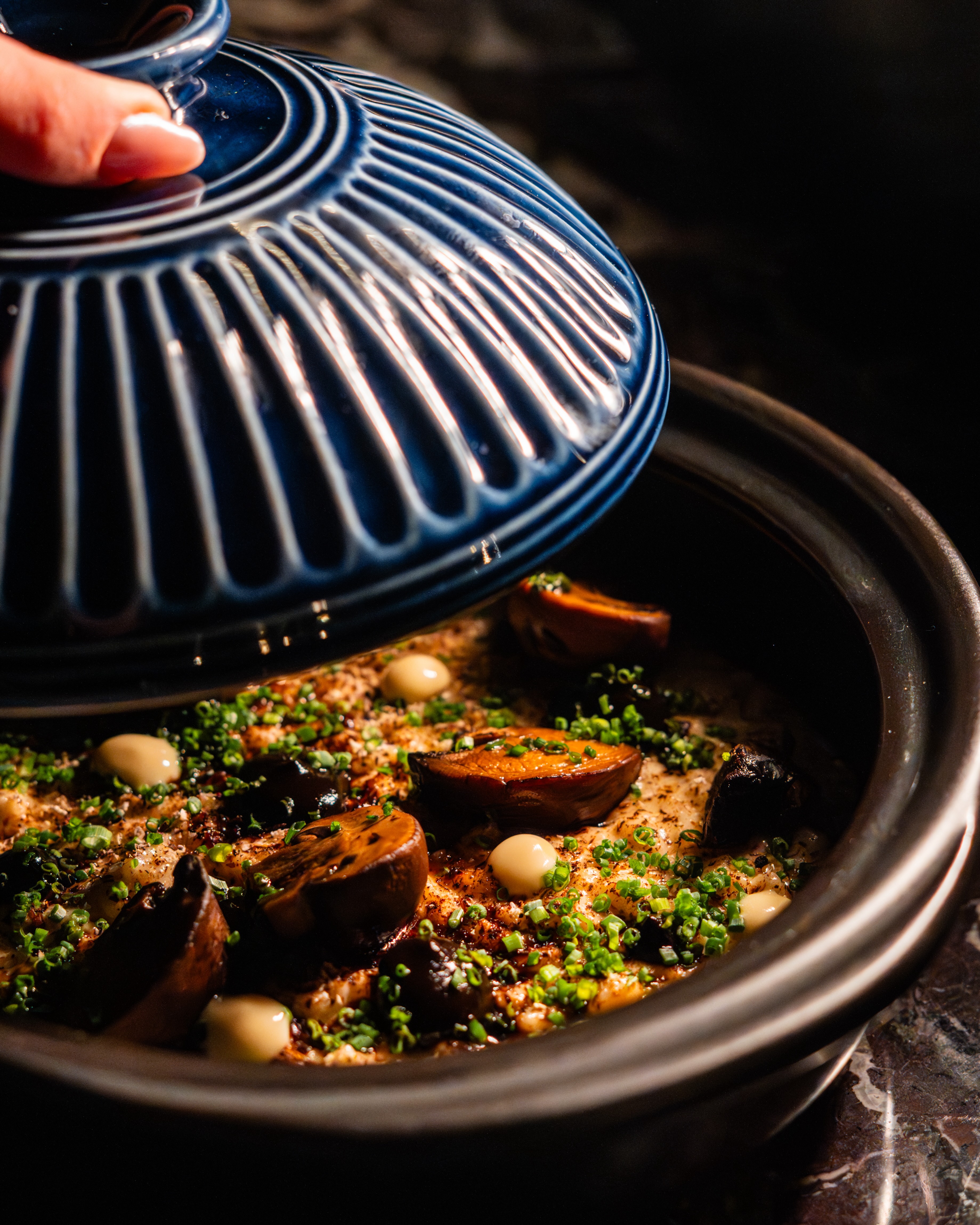 A close-up of a blue-lidded ceramic pot being opened to reveal a savory dish topped with sliced mushrooms, dollops of cream, and freshly chopped chives.