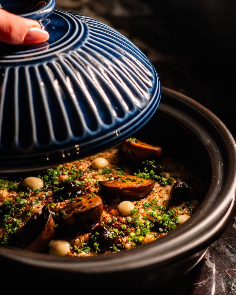 A close-up of a blue-lidded ceramic pot being opened to reveal a savory dish topped with sliced mushrooms, dollops of cream, and freshly chopped chives.