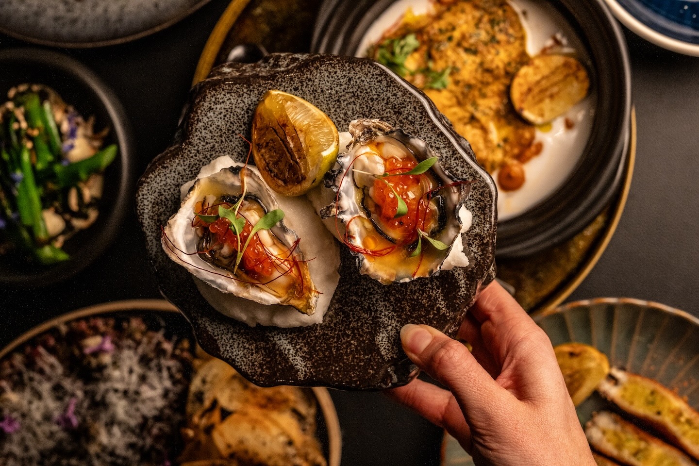 An overhead view of a person's hand holding a small textured plate with two fresh oysters topped with salmon roe and micro-greens. Other small plates including grilled lemon and various appetizers are visible on the dark table below.